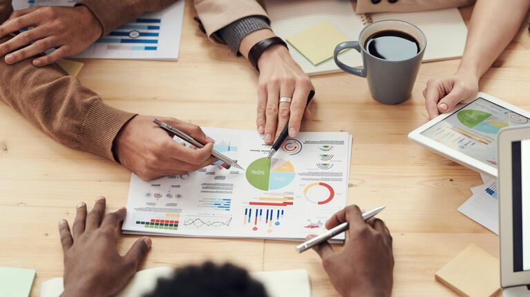 Die Hände mehrerer Personen analysieren ein Arbeitsblatt mit mehreren Tabellen und Diagrammen